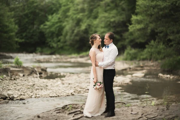 Bellissimo matrimonio coppia baciare e abbracciare vicino alla riva di un fiume di montagna con pietre — Foto Stock