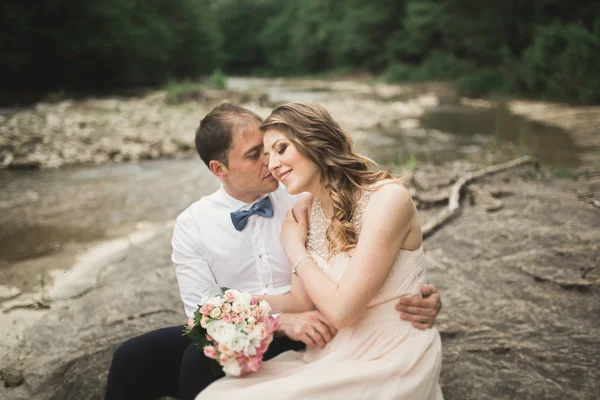 Elegante novio elegante y elegante novia cerca del río con piedras. Pareja de boda enamorada —  Fotos de Stock