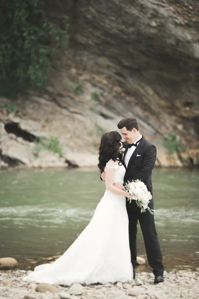 Schönes Hochzeitspaar küsst und umarmt sich am Ufer eines Gebirgsflusses mit Steinen — Stockfoto