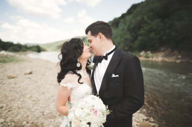 Güzel düğün çifti bir dağ nehrinin kıyısında taşlarla öpüşüp kucaklaşıyorlar.