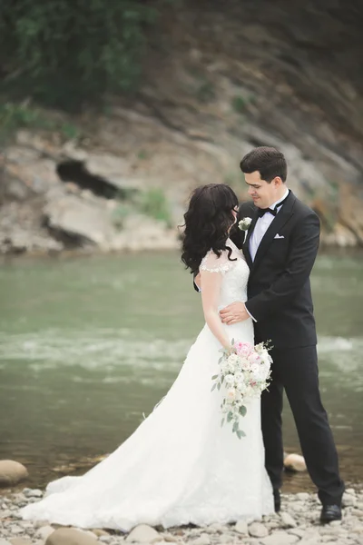 Elegante elegante noiva morena feliz e lindo noivo no fundo de uma bela cachoeira nas montanhas — Fotografia de Stock