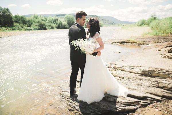 Elegante stilvolle glückliche brünette Braut und wunderschöner Bräutigam vor dem Hintergrund eines wunderschönen Wasserfalls in den Bergen — Stockfoto