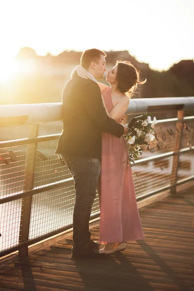 Elegáns szerelmes esküvő házaspár, vőlegény, menyasszony, rózsaszín ruhát csókolózás, és átölelte a hídon naplementekor — Stock Fotó