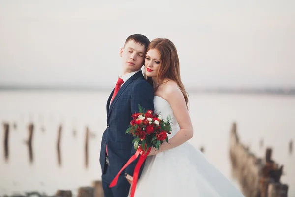 Wedding couple, groom, bride with bouquet posing near sea on sunset — Stock Photo, Image