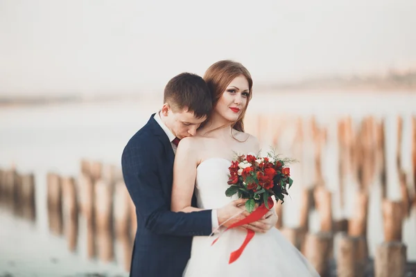 Piękny ślub młodej pary, Narzeczeni pozowanie w pobliżu drewnianych słupów na tle morza — Zdjęcie stockowe