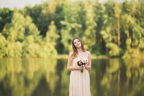 Великолепная невеста в элегантном платье держа букет, позируя возле леса и озера — стоковое фото