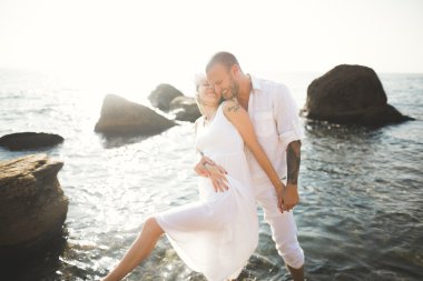 Düğün çifti mavi denizin yanındaki kayalarda öpüşüyor ve sarılıyor.