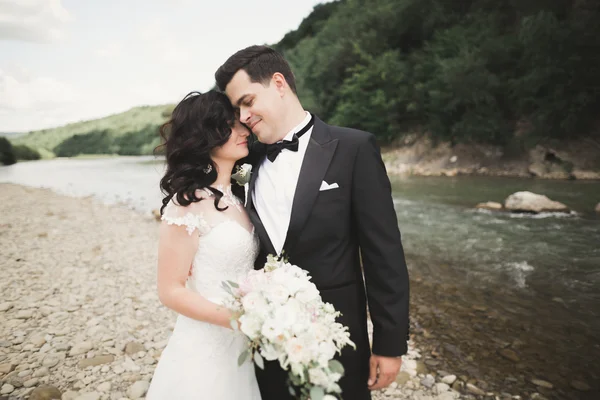 Elegante stilvolle glückliche brünette Braut und wunderschöner Bräutigam vor dem Hintergrund eines wunderschönen Wasserfalls in den Bergen — Stockfoto