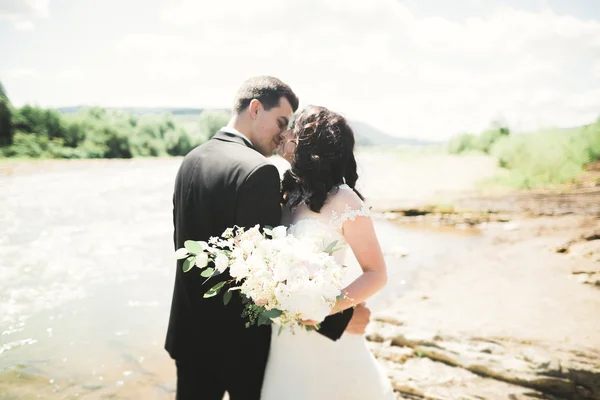 Hermosa pareja de boda besándose y abrazándose cerca de la orilla de un río de montaña con piedras —  Fotos de Stock