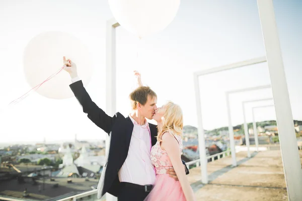 Retrato ao ar livre muito ensolarado de jovem casal elegante enquanto se beija no telhado com vista para a cidade — Fotografia de Stock