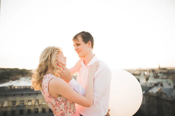 Jovem casal apaixonado posando no telhado com vista perfeita da cidade de mãos dadas e abraços. Belo pôr do sol — Fotografia de Stock