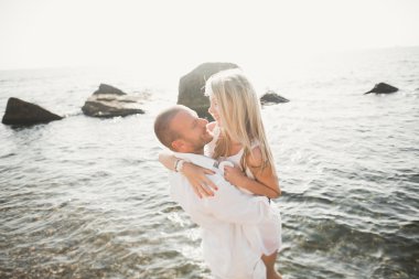 Düğün çifti mavi denizin yanındaki kayalarda öpüşüyor ve sarılıyor.