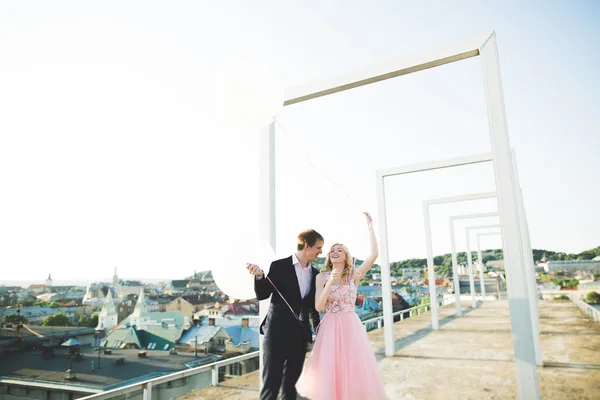 Retrato ao ar livre muito ensolarado de jovem casal elegante enquanto se beija no telhado com vista para a cidade — Fotografia de Stock