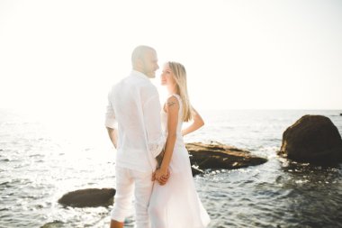 Düğün çifti mavi denizin yanındaki kayalarda öpüşüyor ve sarılıyor.