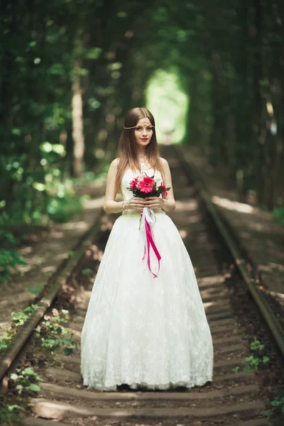 Hermosa novia morena en elegante vestido blanco sosteniendo ramo posando árboles limpios — Foto de Stock