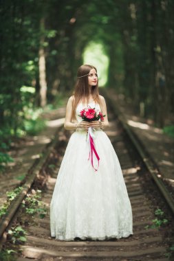 Zarif beyaz elbiseli güzel esmer gelin elinde buket ile güzel ağaçlar poz veriyor.