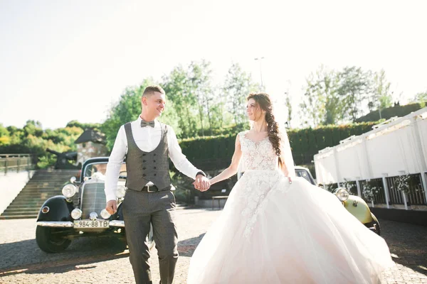 Pareja de boda, novia hermosa y novio elegante tomados de la mano y mirándose el uno al otro cerca de coche de boda retro — Foto de Stock