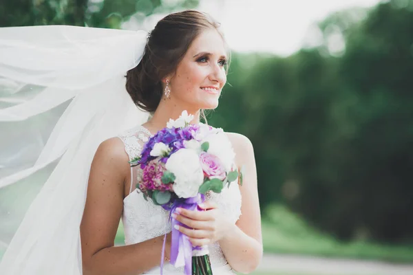 Hermosa novia en elegante vestido blanco sosteniendo ramo posando en el parque — Foto de Stock