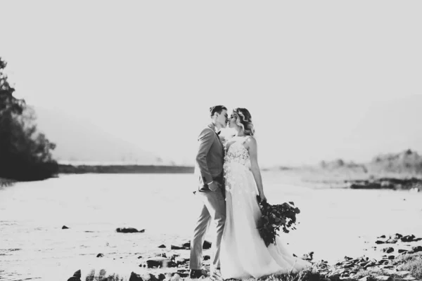 Hermosa pareja de boda besándose y abrazándose cerca de la orilla de un río de montaña con piedras —  Fotos de Stock