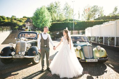 Düğün çifti, güzel gelin ve zarif damat el ele tutuşup birbirlerine retro düğün arabasının yanında bakıyorlar.