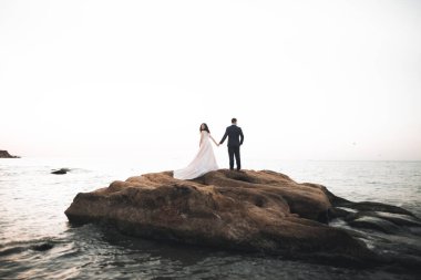 Düğün çifti mavi denizin yanındaki kayalarda öpüşüyor ve sarılıyor.