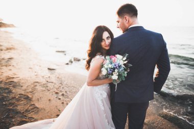 Düğün çifti mavi denizin yanındaki kayalarda öpüşüyor ve sarılıyor.