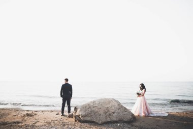 Düğün çifti mavi denizin yanındaki kayalarda öpüşüyor ve sarılıyor.