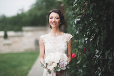 Zarif beyaz elbiseli güzel esmer gelin elinde buket ile güzel ağaçlar poz veriyor.