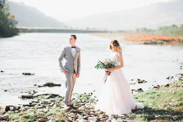 Hermosa pareja de boda besándose y abrazándose cerca de la orilla de un río de montaña con piedras —  Fotos de Stock