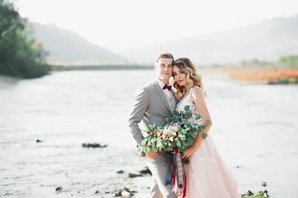 Hermosa pareja de boda besándose y abrazándose cerca de la orilla de un río de montaña con piedras — Foto de Stock