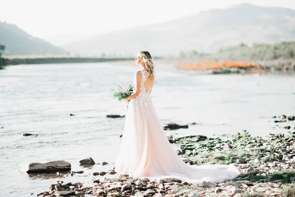 Bella sposa bruna in elegante vestito bianco che tiene bouquet in posa vicino al fiume — Foto Stock