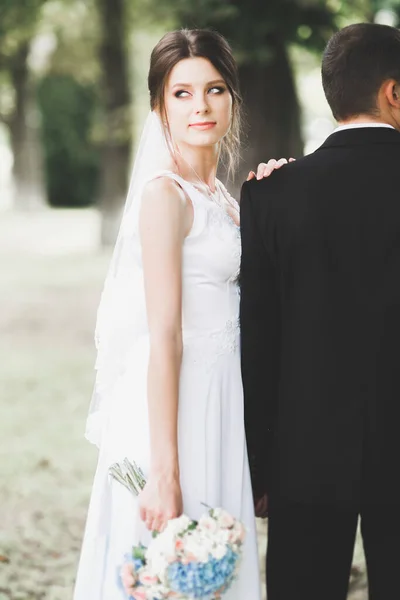 Elegante coppia di sposi felici che passeggiano nel parco il giorno del loro matrimonio con bouquet — Foto Stock