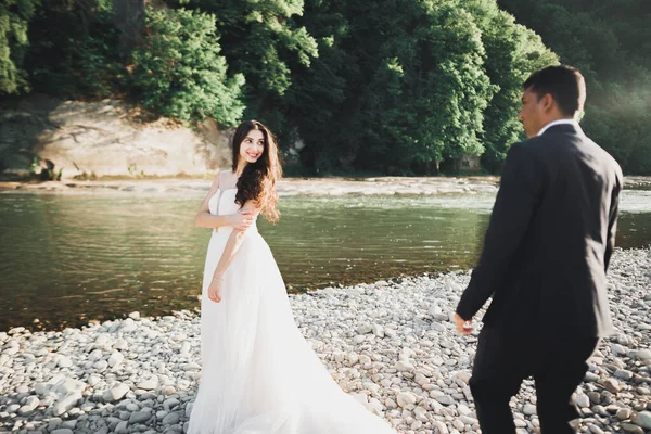 Elegante novia rubia feliz y elegante novio en el fondo de un hermoso río en las montañas —  Fotos de Stock