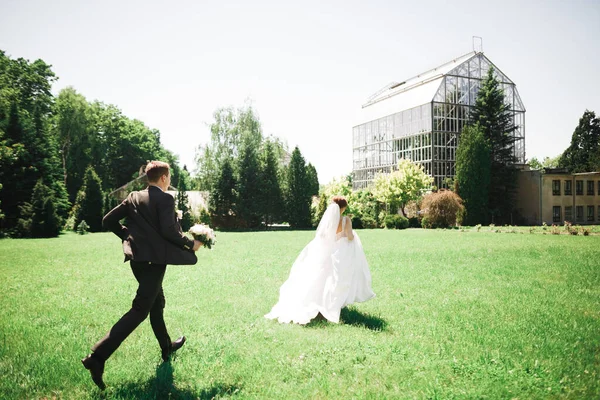 Stilvolles Paar glücklicher Frischvermählter. Braut läuft dem Bräutigam am Hochzeitstag mit Blumenstrauß im Park davon — Stockfoto