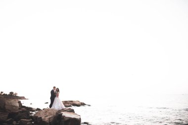 Düğün çifti mavi denizin yanındaki kayalarda öpüşüyor ve sarılıyor.