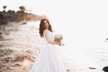 Güzel bayan, gelin gün batımında deniz kenarında bir gelinlik içinde poz veriyor.