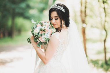 Zarif beyaz elbiseli güzel esmer gelin elinde buket ile güzel ağaçlar poz veriyor.