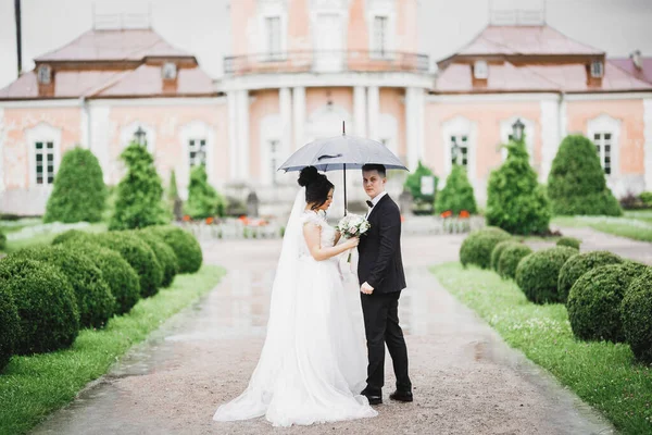 Schöne junge Hochzeitspaar posiert mit Blumenstrauß in den Händen unter Regenschirm, während es regnet — Stockfoto