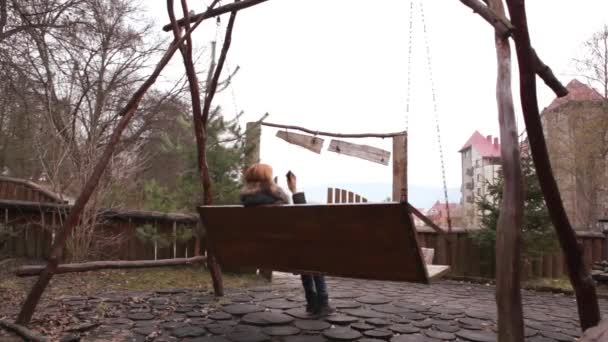 Дівчина гойдається на дерев'яній гойдалці і розмовляє по телефону — стокове відео