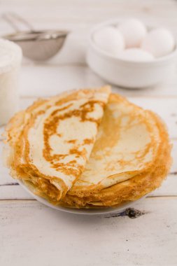 İnce, leziz yuvarlak krepler beyaz bir tabakta, ahşap masada, mutfakta yumurta ve un ile.
