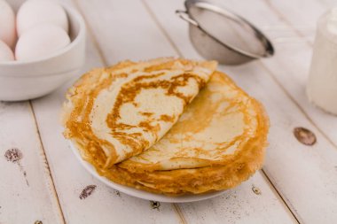 İnce, leziz yuvarlak krepler beyaz bir tabakta, ahşap masada, mutfakta yumurta ve un ile.