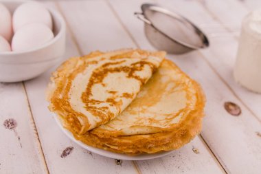 İnce, leziz yuvarlak krepler beyaz bir tabakta, ahşap masada, mutfakta yumurta ve un ile.