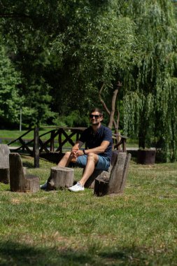 Koyu renk saçlı ve gözlüklü bir adam parkta bir kütüğe oturur ve yazın şort giyer.