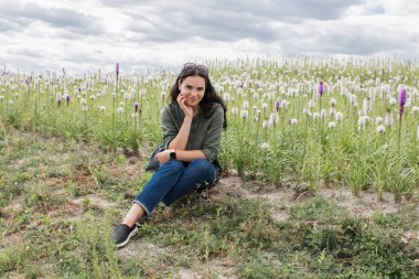 Koyu saçlı bir kız açık bir yaz gününde elinde çiçeklerle tarlada yürür.