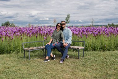 Bir adam ve aşık bir kız çiçeklerin yanındaki bankta oturuyorlar.