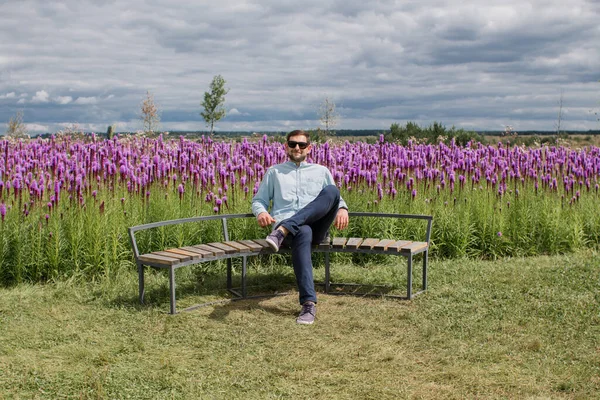 guy in a shirt and pants sits on a bench in a park near a field of flowers