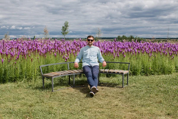 guy in a shirt and pants sits on a bench in a park near a field of flowers