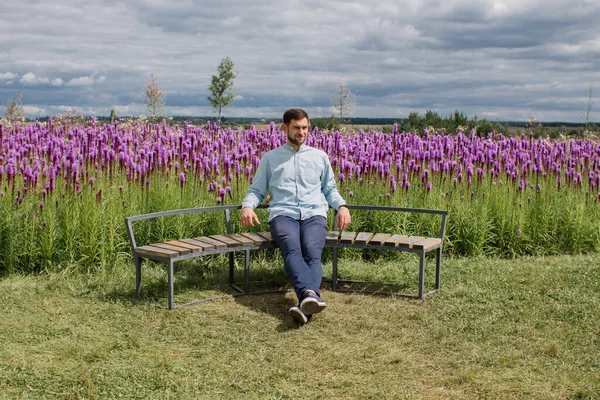 guy in a shirt and pants sits on a bench in a park near a field of flowers