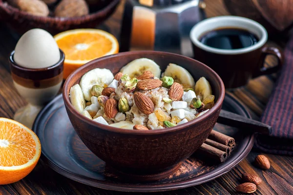 Muz, bal, badem, Antep fıstığı, Hindistan cevizi cips, portakal, tarçın ve kuru üzüm ile karanlık seramik kase organik yulaf lapası. Sağlıklı kahvaltı - sağlık ve diyet konsepti üzerine ahşap masa.