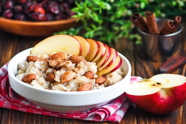 リンゴ、アーモンド、蜂蜜、シナモンと白いセラミックボウルの有機オートミールお粥。健康的な朝食 - 木製のテーブルの上の健康と食事の概念、クローズアップ. — ストック写真